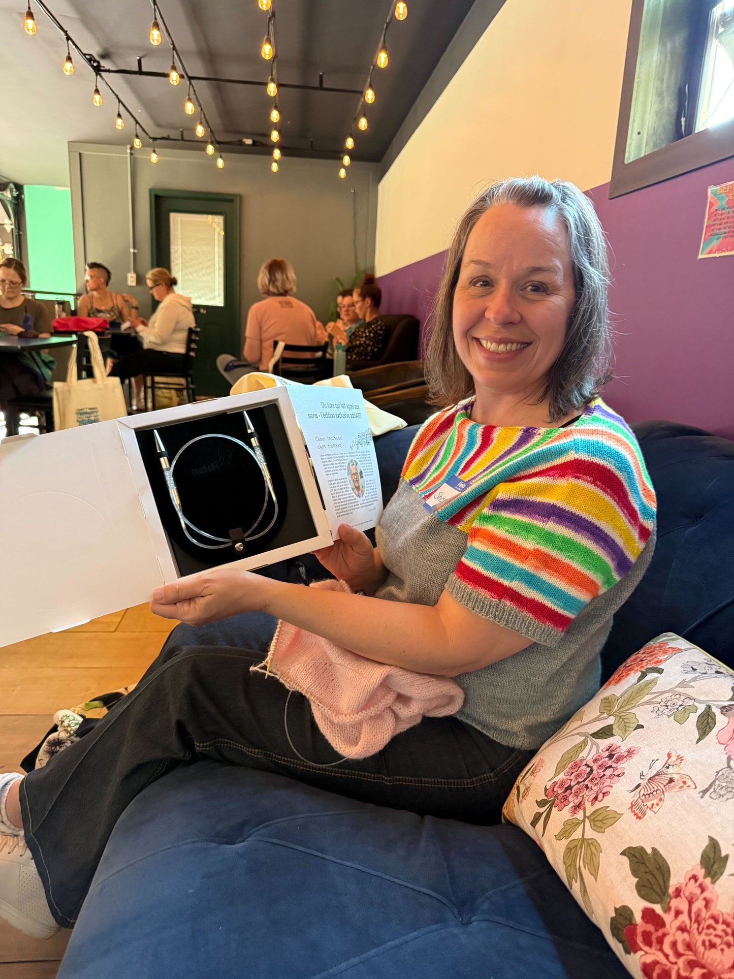 A Stitch ’n Bitch prize winner sitting on a blue couch and holding a box of circular knitting needles, with other makers gathered in the background at Barley Pop Brewing.