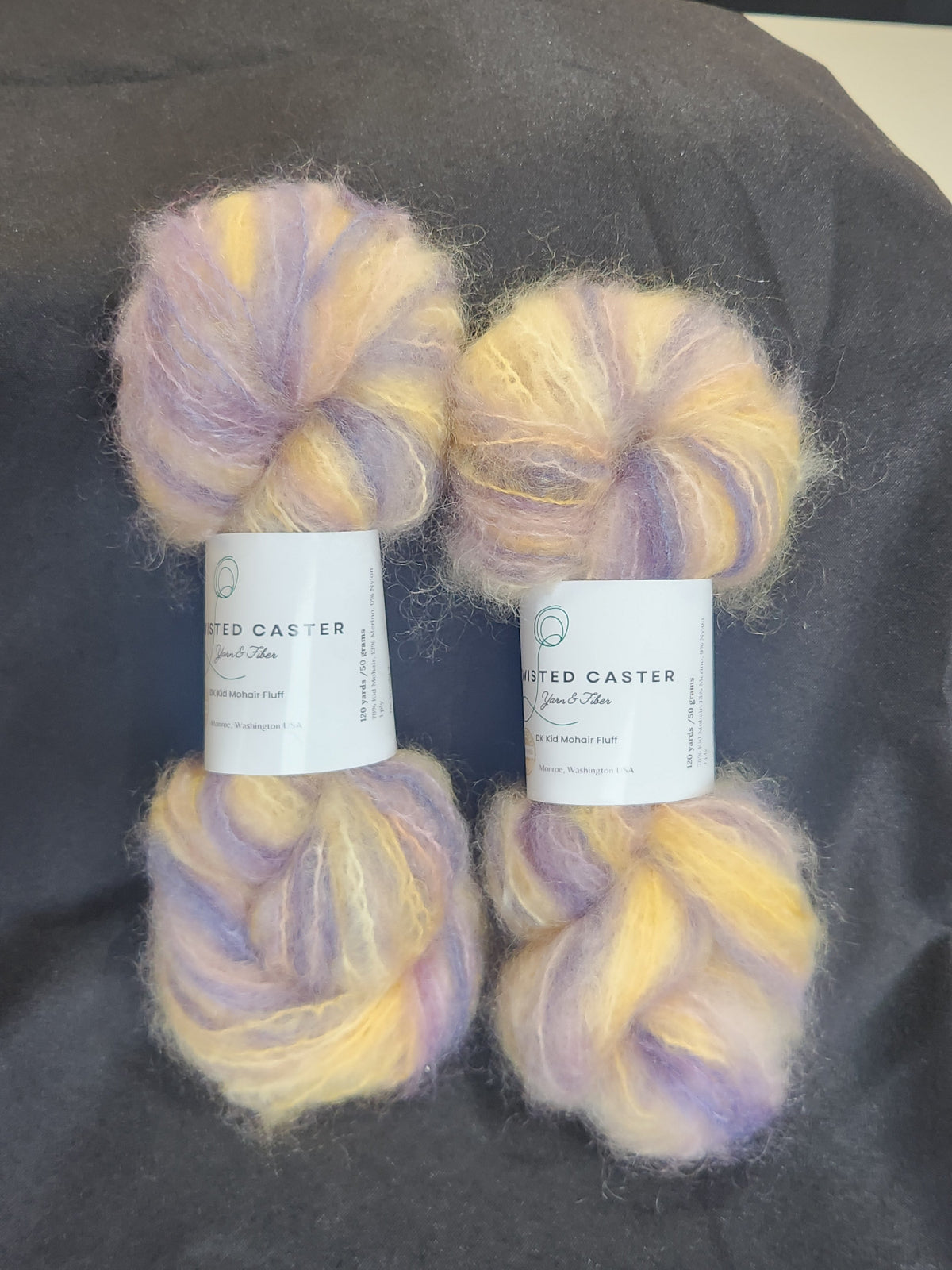 Four skeins of fluffy variegated yarn in Lavender & Yellow, arranged on a dark gray fabric background.