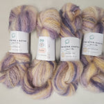 Four skeins of twisted  yarn arranged vertically on a light surface. The yarn has a variegated color pattern of purple, lavender, cream, and pale yellow, with white paper labels in the center.
