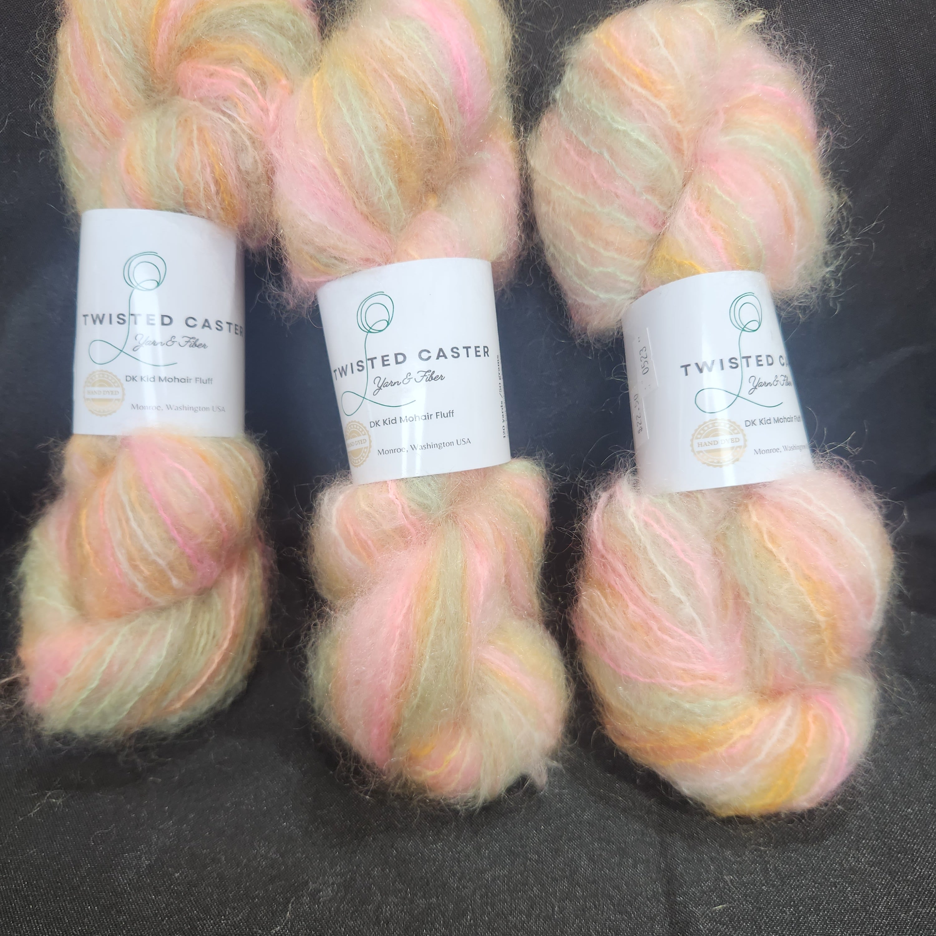 Three skeins of fluffy variegated yarn with white paper labels, featuring soft pastel pink, peach, and pale yellow tones, laid flat on a dark textured background.
