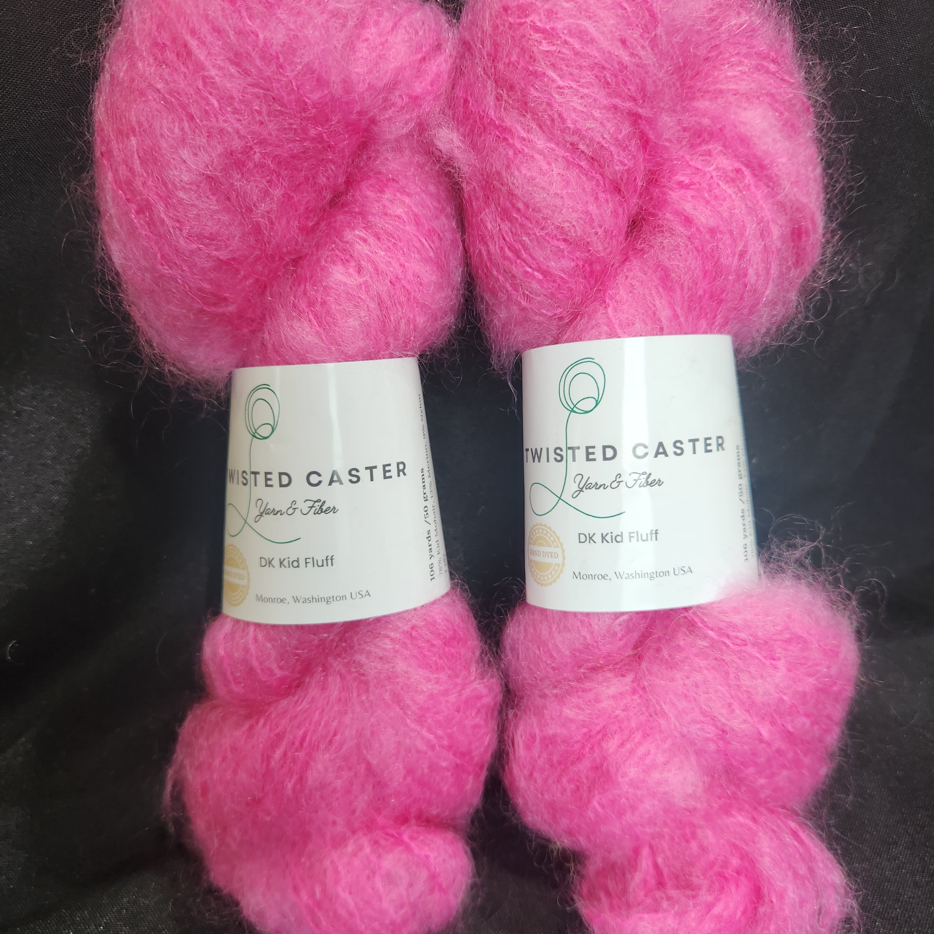 Two fluffy skeins of bright pink yarn with white paper labels, arranged on a dark textured background.