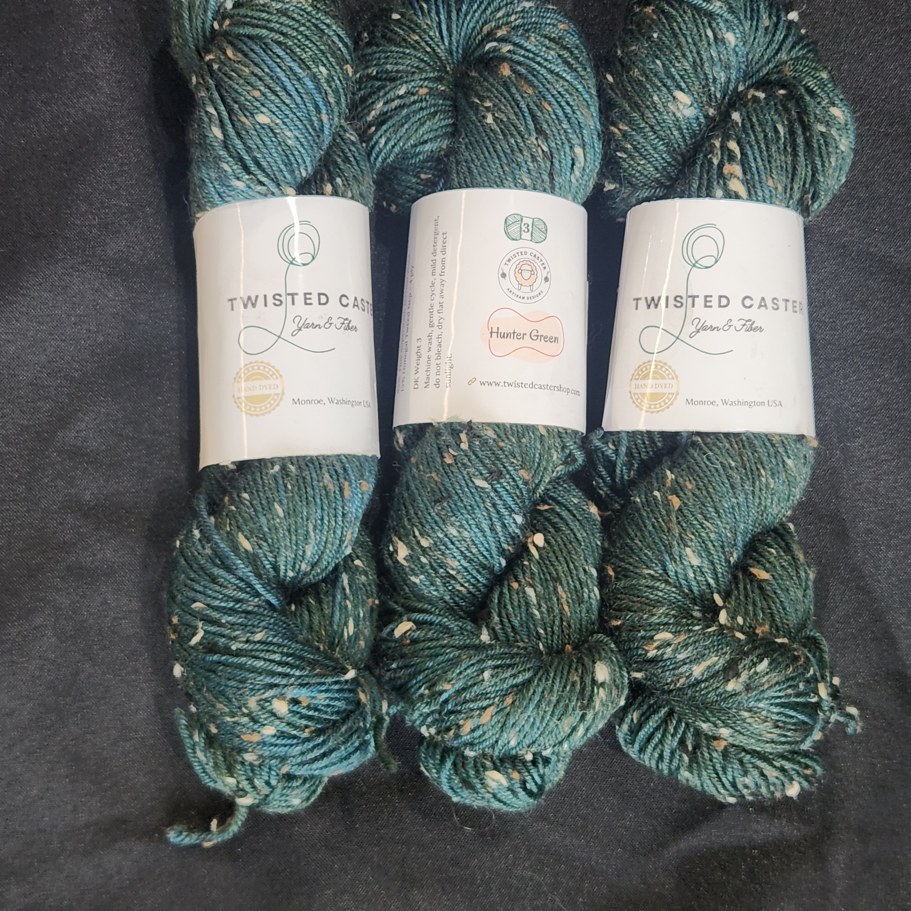 Three skeins of twisted cotton yarn in a teal blue-green color with white flecks, arranged vertically on a dark gray fabric background.