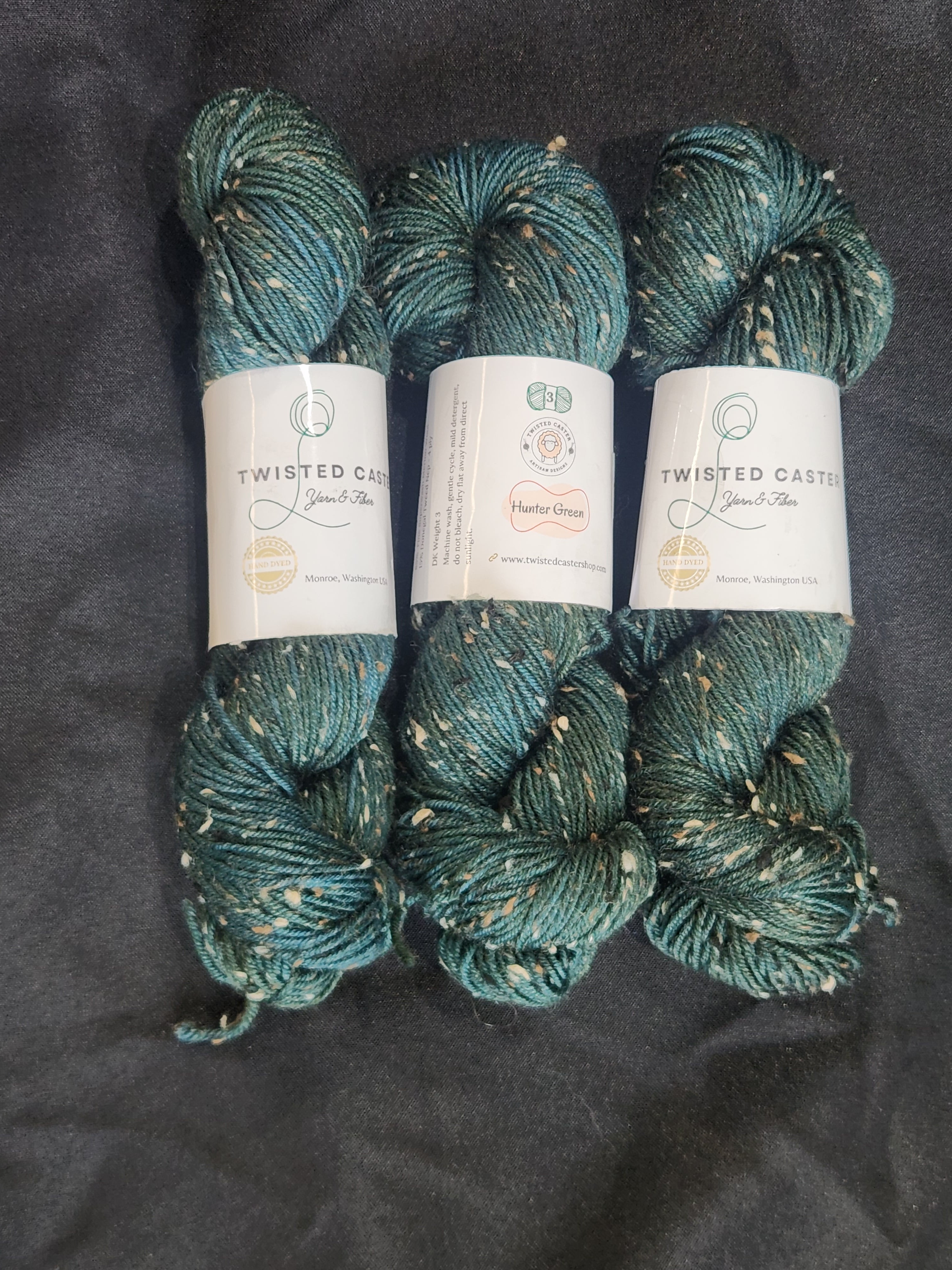 Three skeins of twisted cotton yarn in a teal blue-green color with white flecks, arranged vertically on a dark gray fabric background.