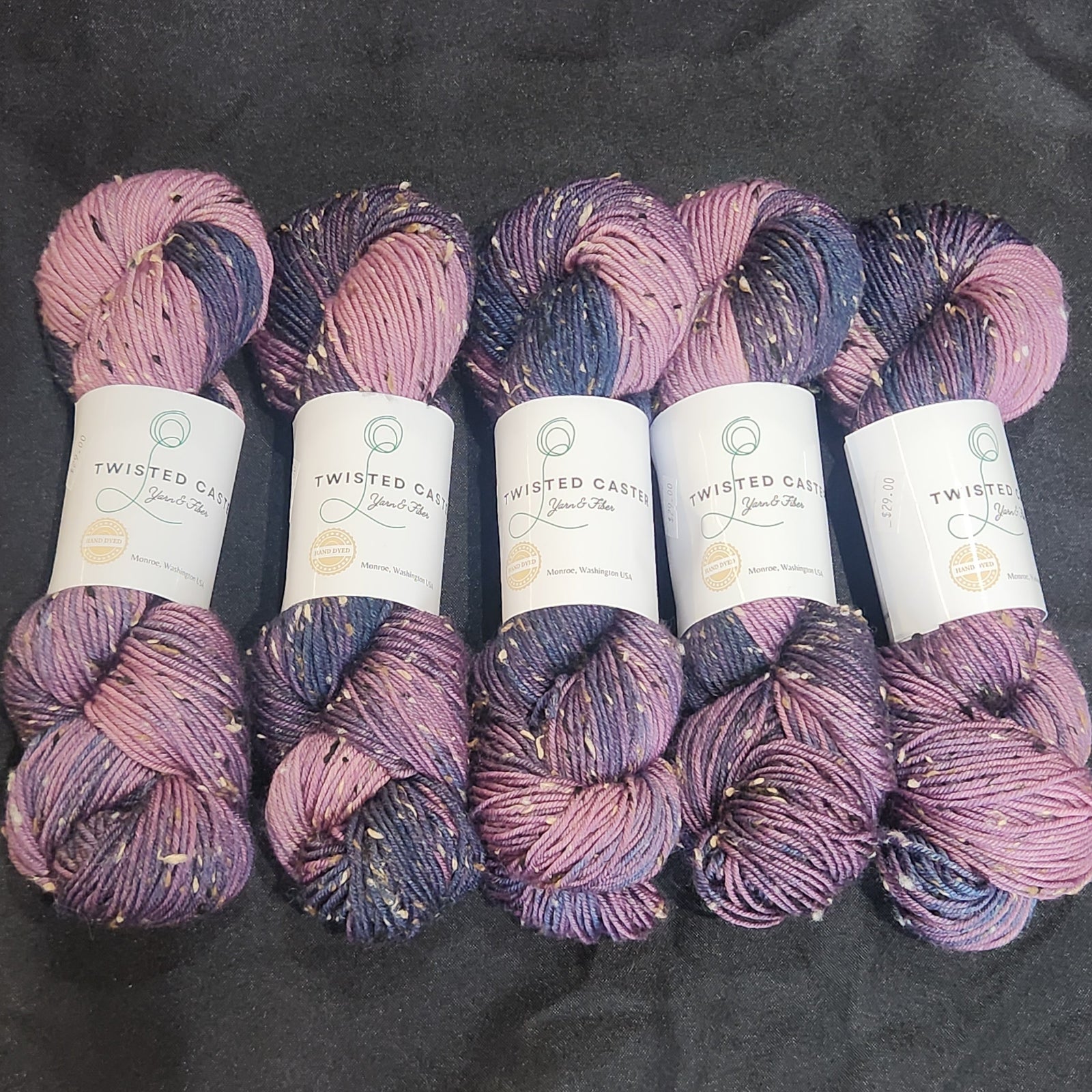 Five skeins of variegated yarn in shades of purple, pink, and navy blue, arranged vertically on a black fabric background. 