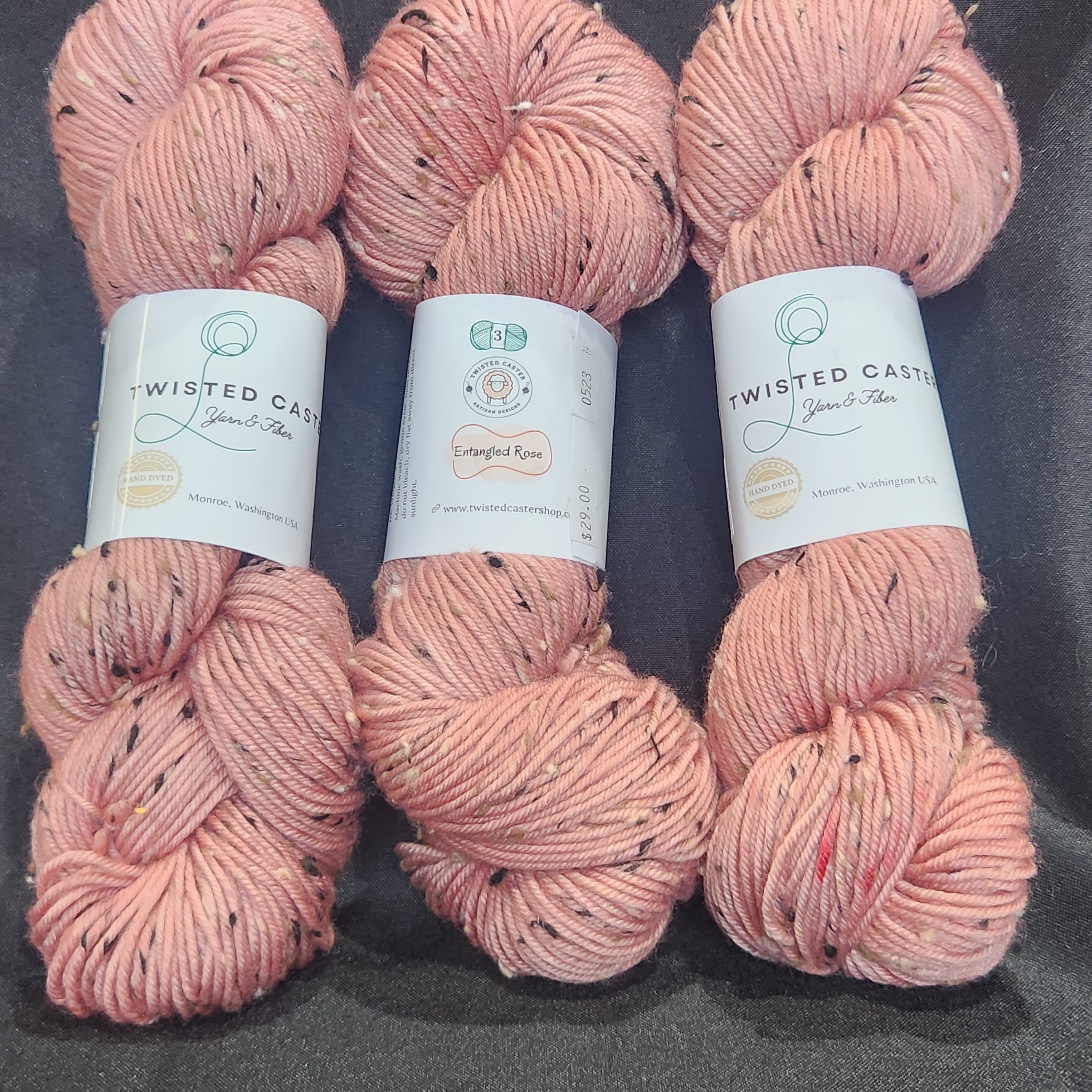 Three skeins yarn in a dusty pink color with small black flecks, laid out horizontally on a dark gray textured background.