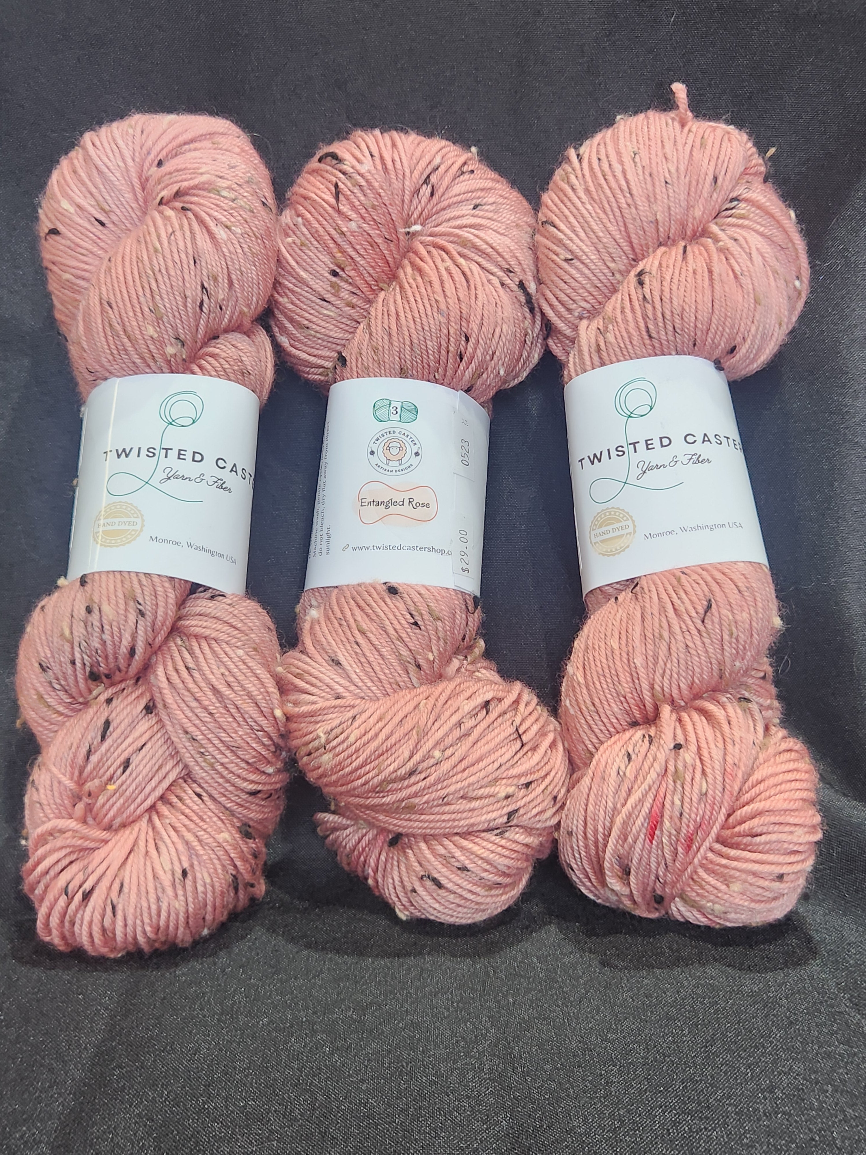 Three skeins yarn in a dusty pink color with small black flecks, laid out horizontally on a dark gray textured background.