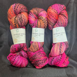 Three skeins of variegated yarn laid side by side on a dark gray fabric background. The yarn features a multicolor variegated pattern with bright pink, orange, purple, and black tones.