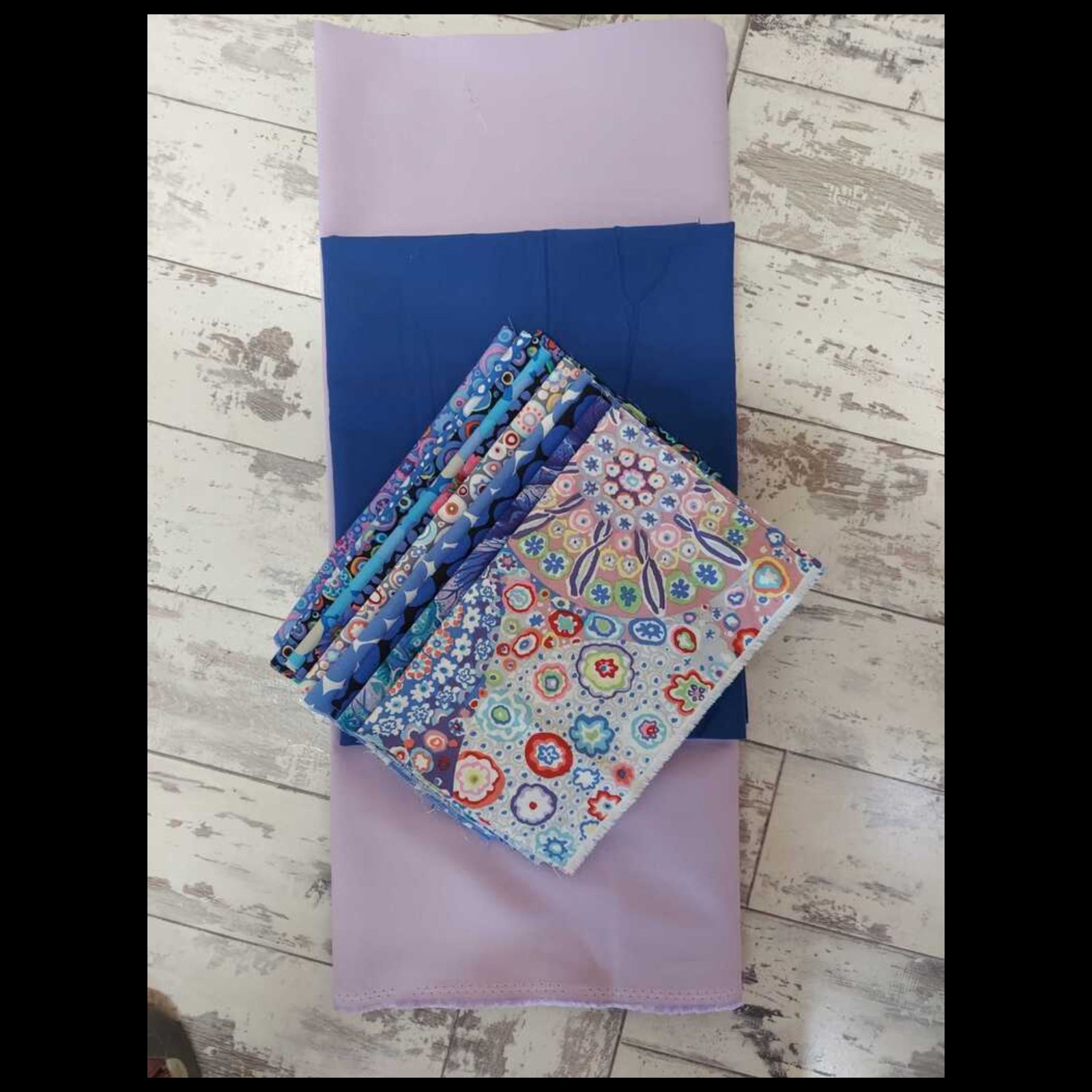 A stack of folded patterned fabric bolts and a larger lavender fabric roll laid on a distressed white wood floor. The fabrics feature various colorful floral, paisley, and geometric patterns, with one patterned fabric showing blue, red, yellow, and green motifs on a light background.