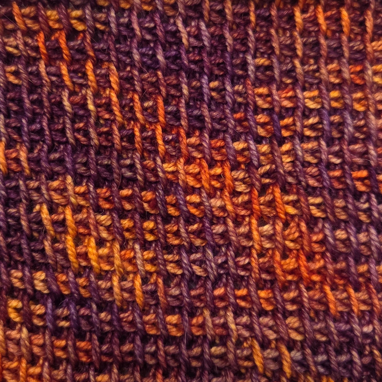 A close-up of a Tunisian crochet fabric swatch, showing a variegated pattern of purple, orange, and brown yarn.