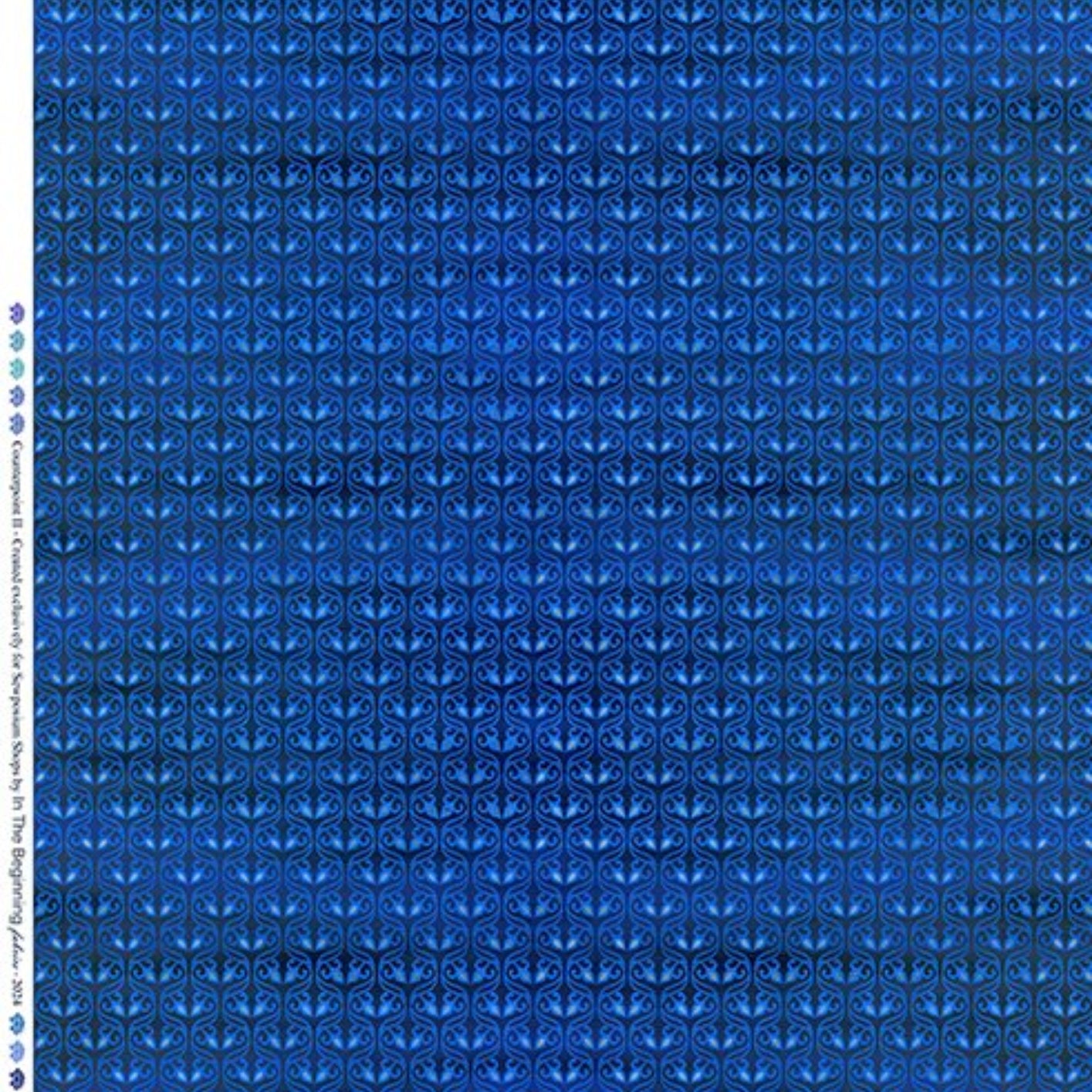A close-up of a blue patterned fabric with a repeating geometric and floral-like motif, shown flat with a ruler along the top edge and a color/brand label along the left side.