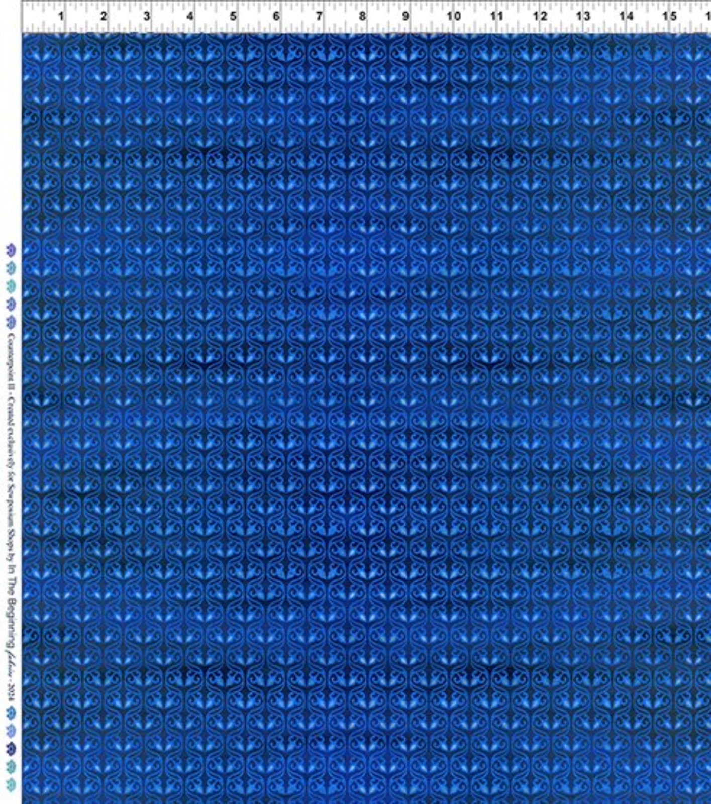 A close-up of a blue patterned fabric with a repeating geometric and floral-like motif, shown flat with a ruler along the top edge and a color/brand label along the left side.