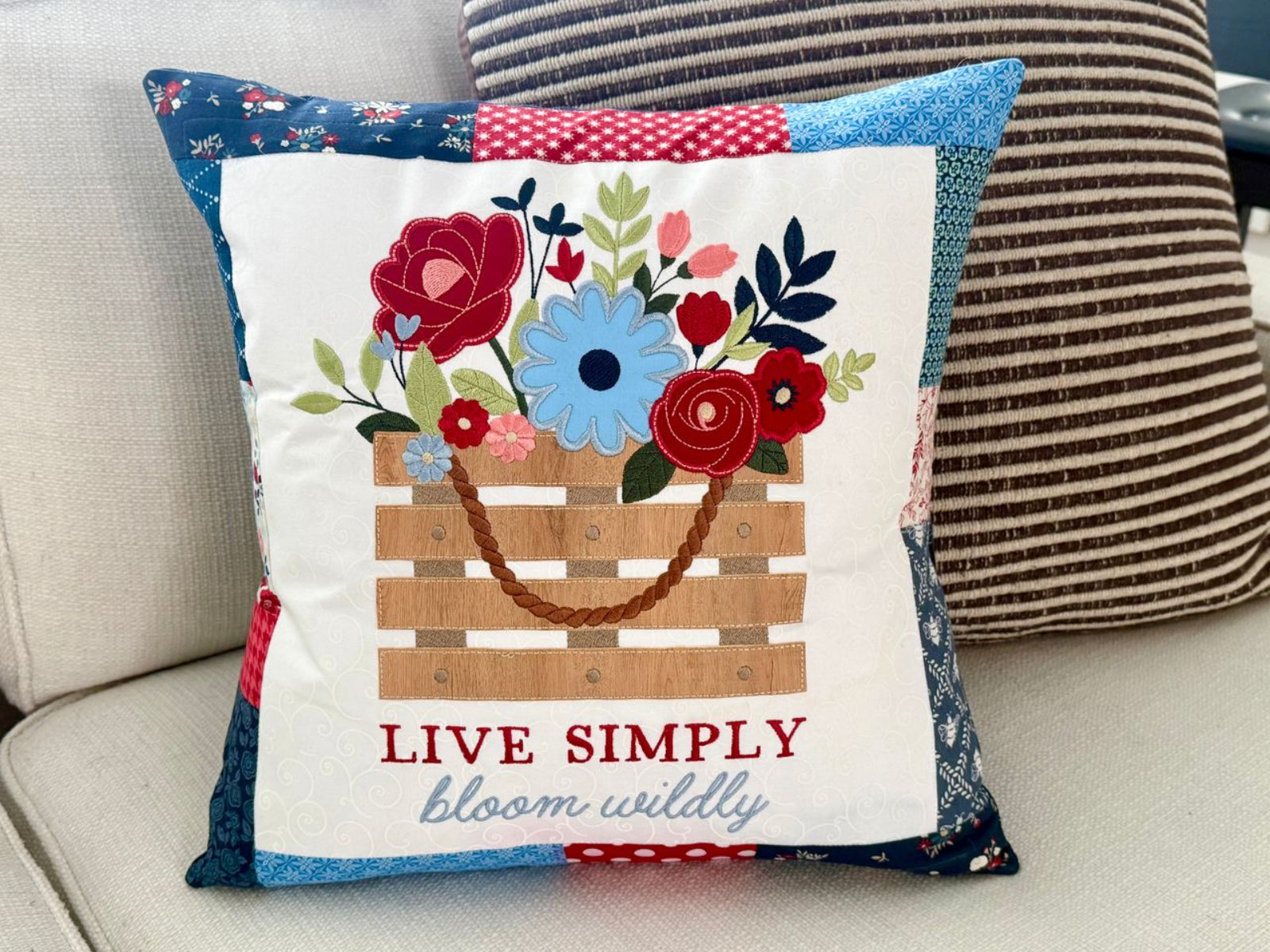 A square decorative throw pillow with a patchwork fabric border and a central applique design of a flower basket. The pillow features the text 'LIVE SIMPLY bloom wildly' and is displayed on a light-colored sofa next to another striped pillow.