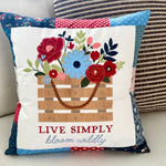 A square decorative throw pillow with a patchwork fabric border and a central applique design of a flower basket. The pillow features the text 'LIVE SIMPLY bloom wildly' and is displayed on a light-colored sofa next to another striped pillow.
