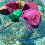 Three‑dimensional embroidered flower with layered pink and yellow petals on a quilted blue‑green background.