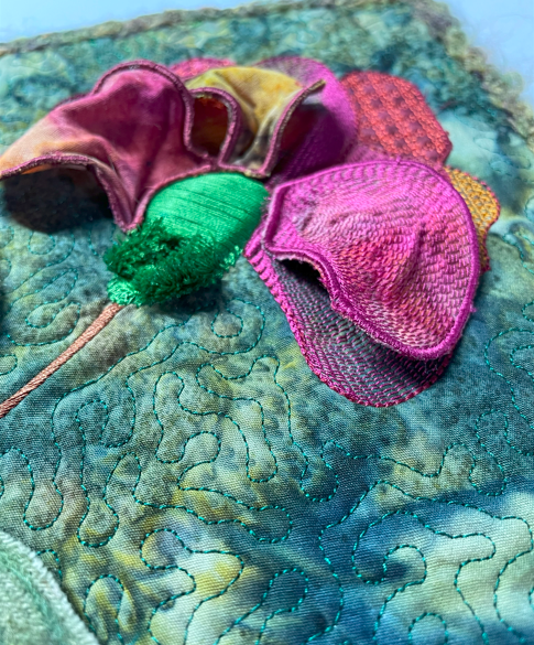 Three‑dimensional embroidered flower with layered pink and yellow petals on a quilted blue‑green background.