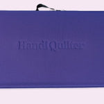 A purple rectangular quilt case with the brand name 'Handi Quilter' embossed in the center, shown against a pale pink background.