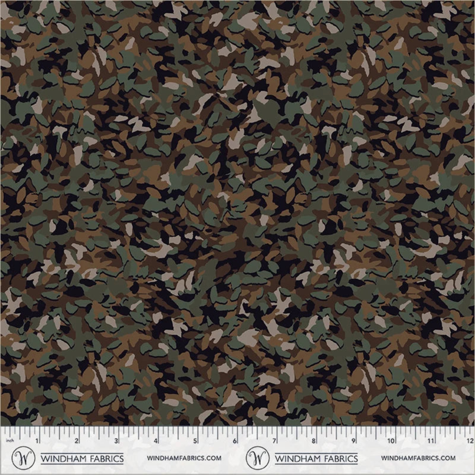 A close-up of a woodland camouflage fabric pattern with green, brown, black, and beige leaf-like shapes, shown with a measuring ruler along the bottom edge from Windham Fabrics.