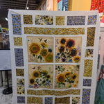 A handmade quilt featuring a sunflower and butterfly design, displayed on a stand inside a craft fair or quilt shop.