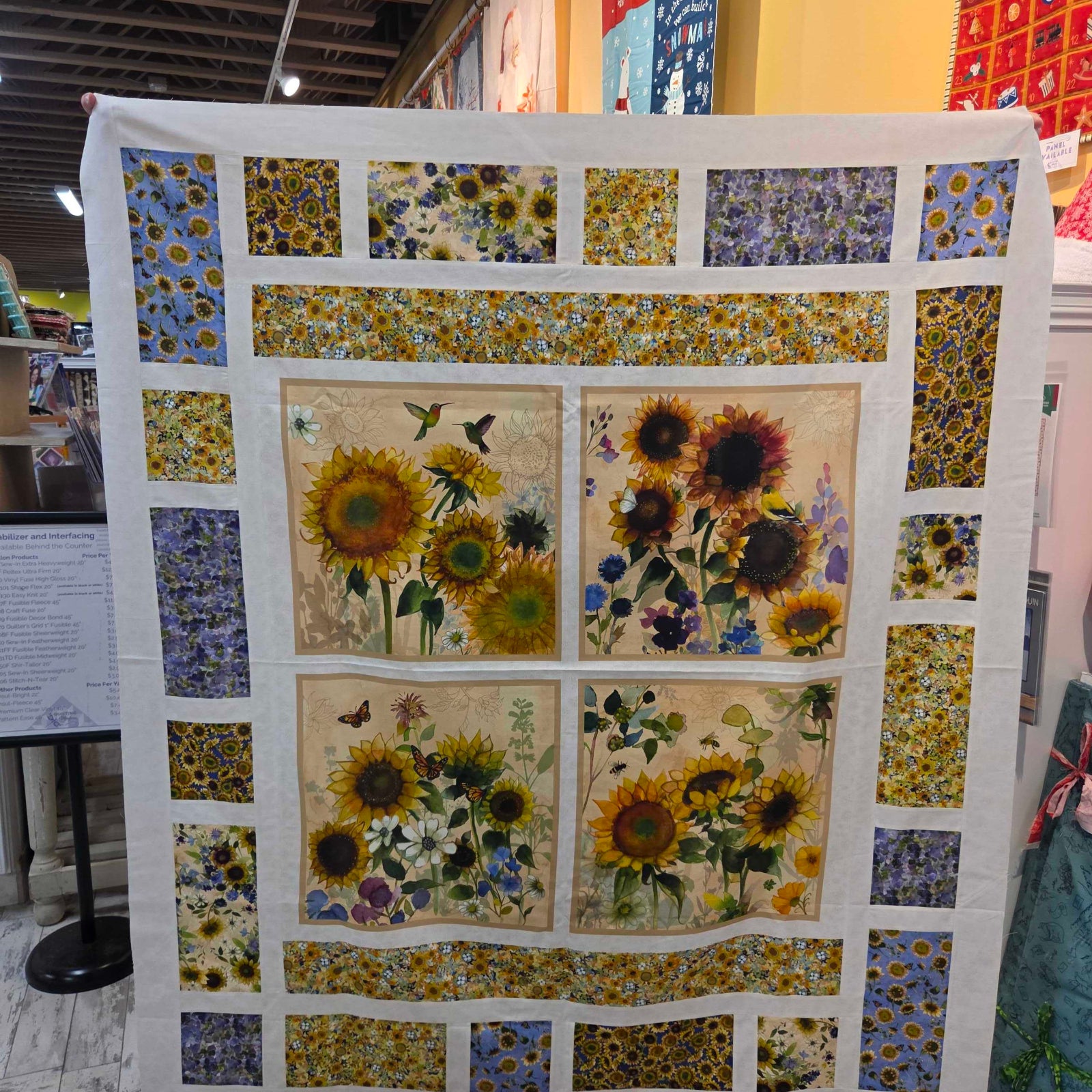 A handmade quilt featuring a sunflower and butterfly design, displayed on a stand inside a craft fair or quilt shop.