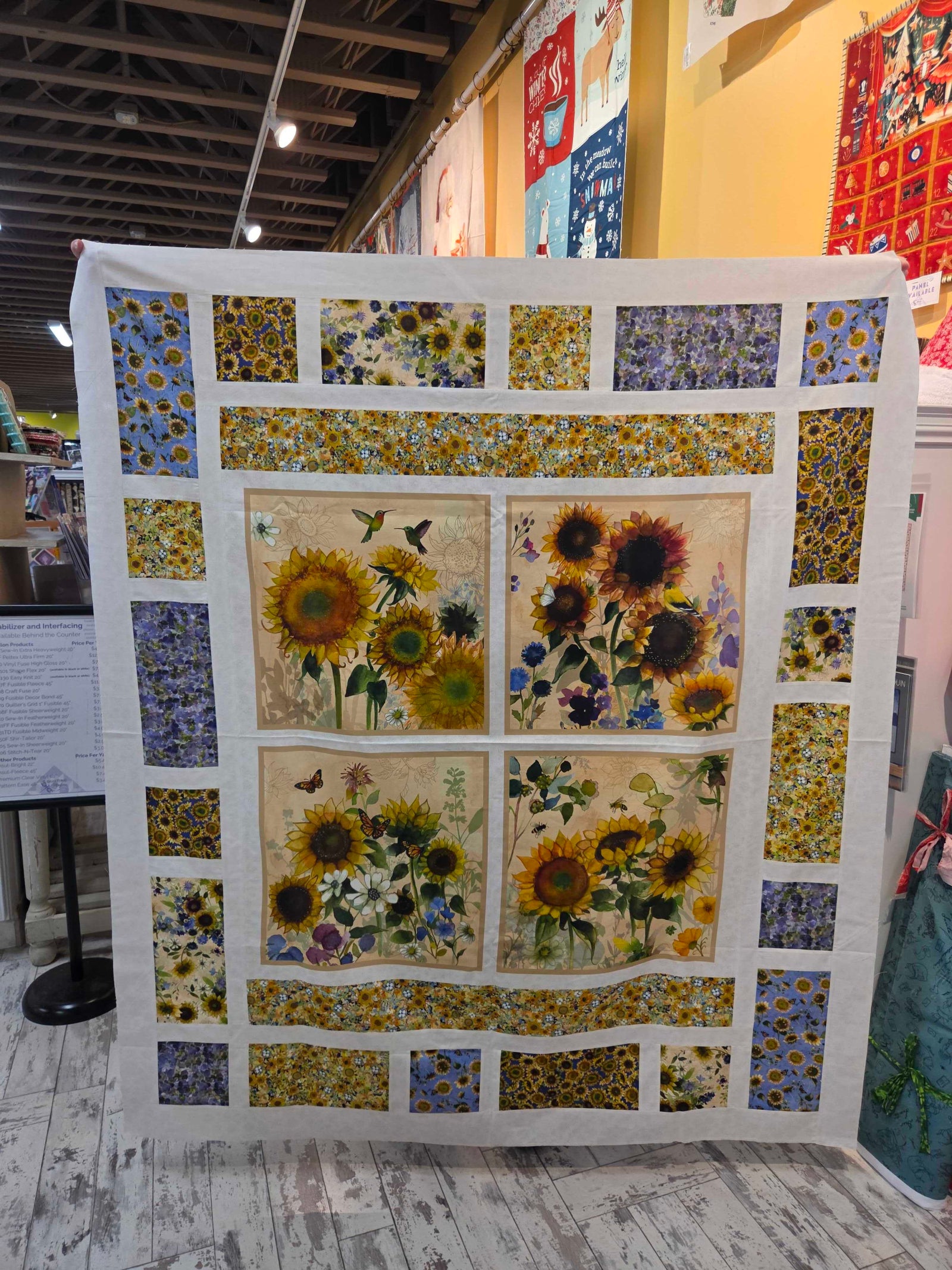 A handmade quilt featuring a sunflower and butterfly design, displayed on a stand inside a craft fair or quilt shop.