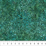 A close-up of a batik fabric with a repeating tropical leaf pattern in shades of green, teal, and turquoise, with a white ruler at the bottom indicating scale.