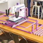 A white and purple sewing machine mounted on a purple metal guide rail system on a wooden table, with matching accessories and small circular pieces arranged beside it. 