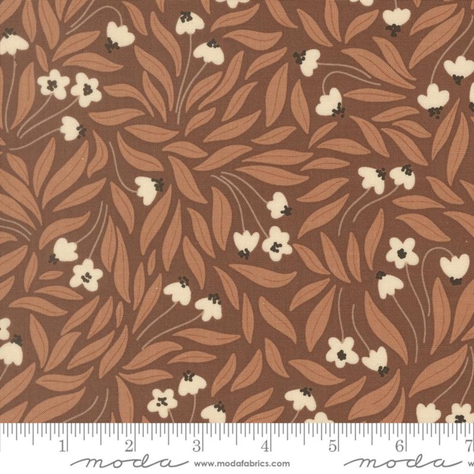 A close-up of a fabric pattern featuring a brown background with a repeating design of light brown leaves and small cream flowers with tiny dark centers. A white ruler scale and the Modas Fabrics logo are visible along the bottom edge.