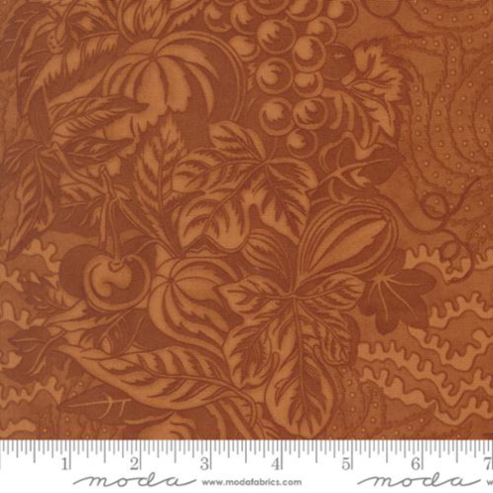 A close-up of a brown fabric with an embossed or printed autumnal pattern featuring pumpkins, gourds, grapes, and leaves. A white ruler along the bottom edge shows scale, and the word 'moda' appears at the bottom.