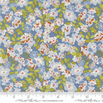A close-up of a floral fabric pattern with white flowers, green leaves, and small orange-red centers on a dusty blue background, with a measuring ruler along the bottom edge and Modas fabric branding at the bottom.