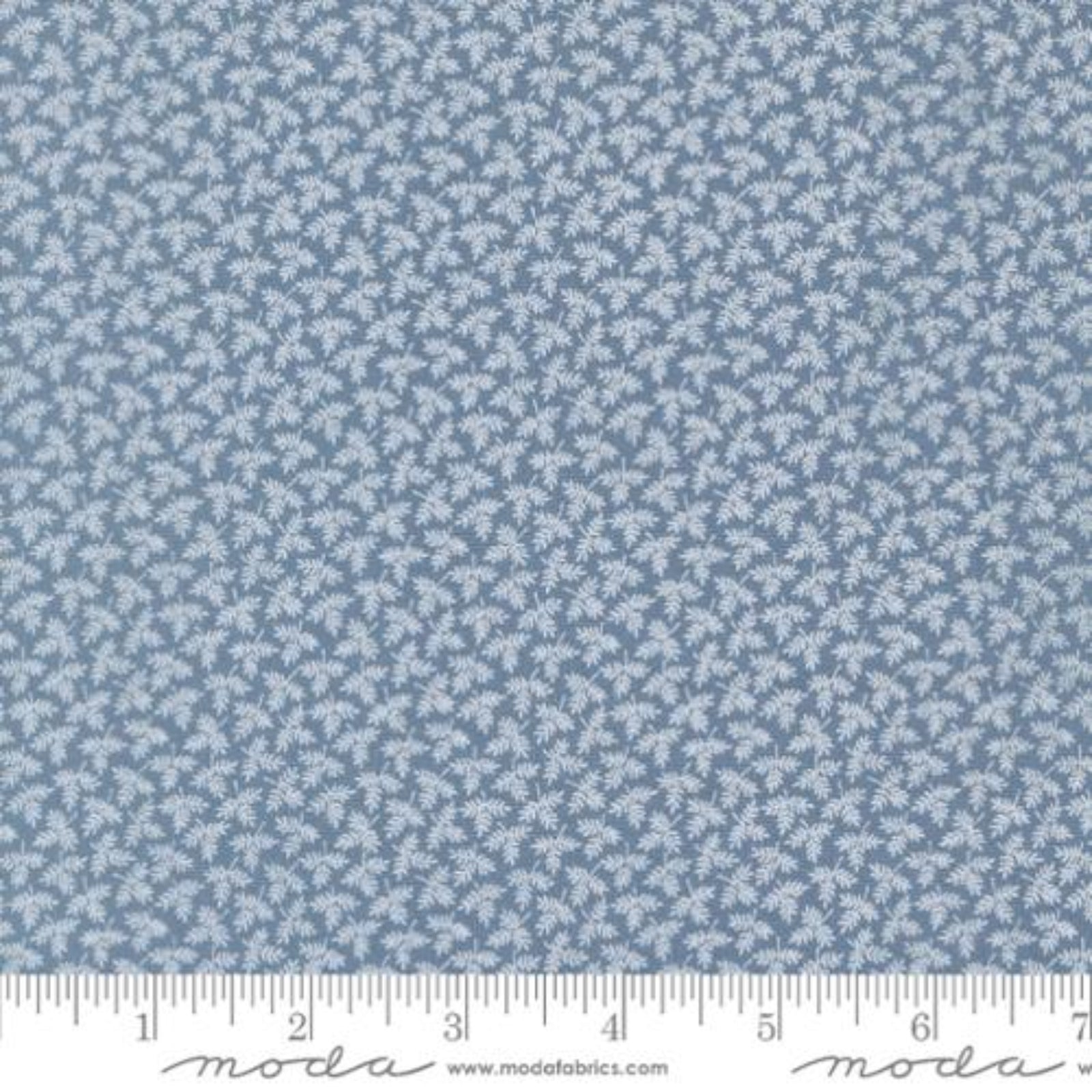 A close-up of a fabric pattern showing a light blue background with a repeating small white floral or leaf-like motif, with a ruler along the bottom edge for scale. The word 'moda' and 'www.