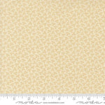 A close-up of a fabric swatch with a repeating small floral or leaf-like pattern in warm beige and cream, with a ruler along the bottom edge showing scale. The word 'moda' and 'www.