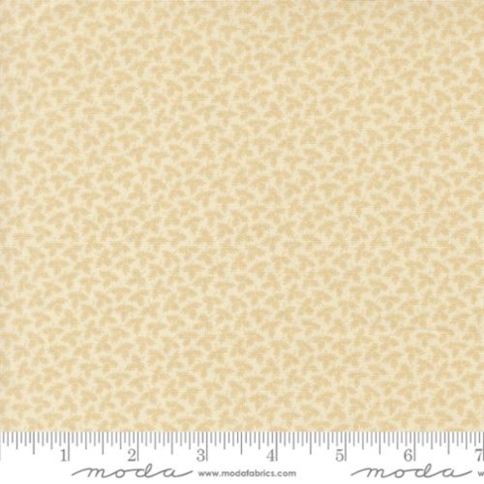 A close-up of a fabric swatch with a repeating small floral or leaf-like pattern in warm beige and cream, with a ruler along the bottom edge showing scale. The word 'moda' and 'www.