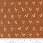 A close-up of a fabric pattern featuring a warm rust-brown background with repeating cream-colored bows and small white dotted motifs, with a ruler along the bottom edge showing scale. The brand name 'moda' is visible on the ruler watermark.