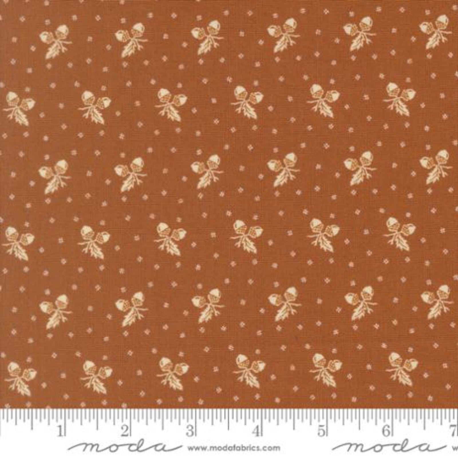 A close-up of a fabric pattern featuring a warm rust-brown background with repeating cream-colored bows and small white dotted motifs, with a ruler along the bottom edge showing scale. The brand name 'moda' is visible on the ruler watermark.