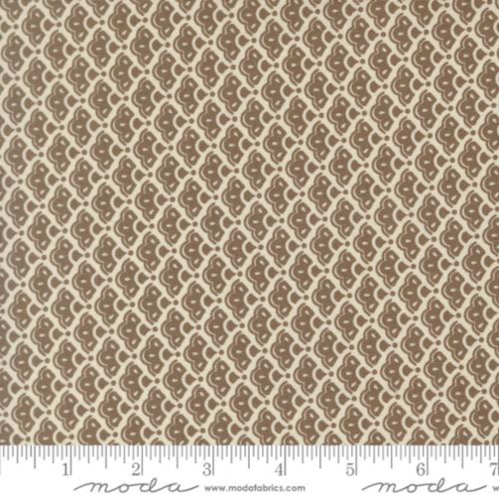 A close-up of a fabric pattern with a repeating brown paisley-like motif on a warm beige background, with a ruler along the bottom edge and Modas Fabrics branding at the bottom.