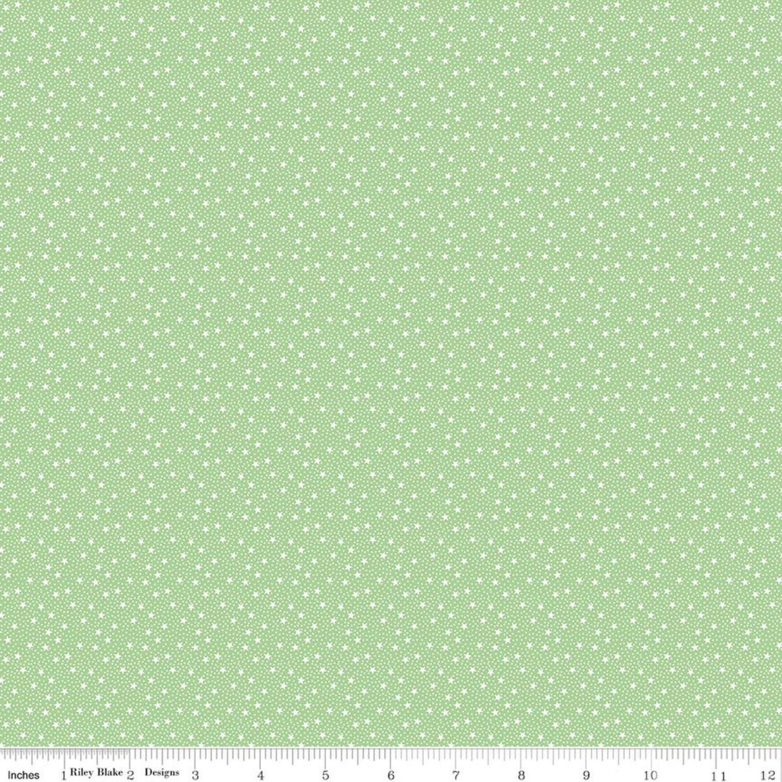 A close-up fabric pattern swatch showing a light sage green background with a repeating white star and ditsy dot motif, with a ruler along the bottom edge for scale.
