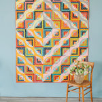 Curvy Log Cabin quilt in bright modern colors displayed on a wall with a chair and flower basket.