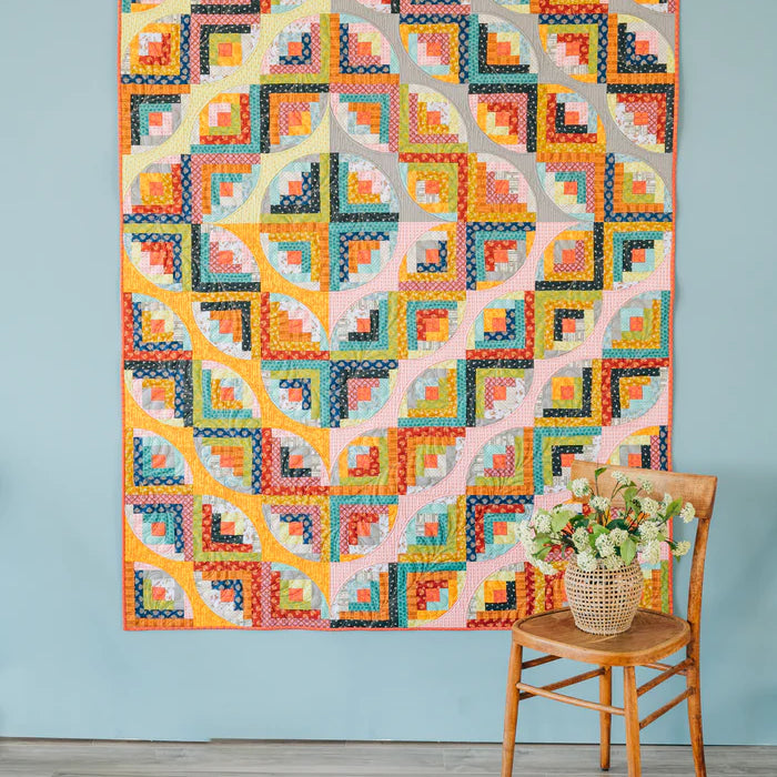 Curvy Log Cabin quilt in bright modern colors displayed on a wall with a chair and flower basket.