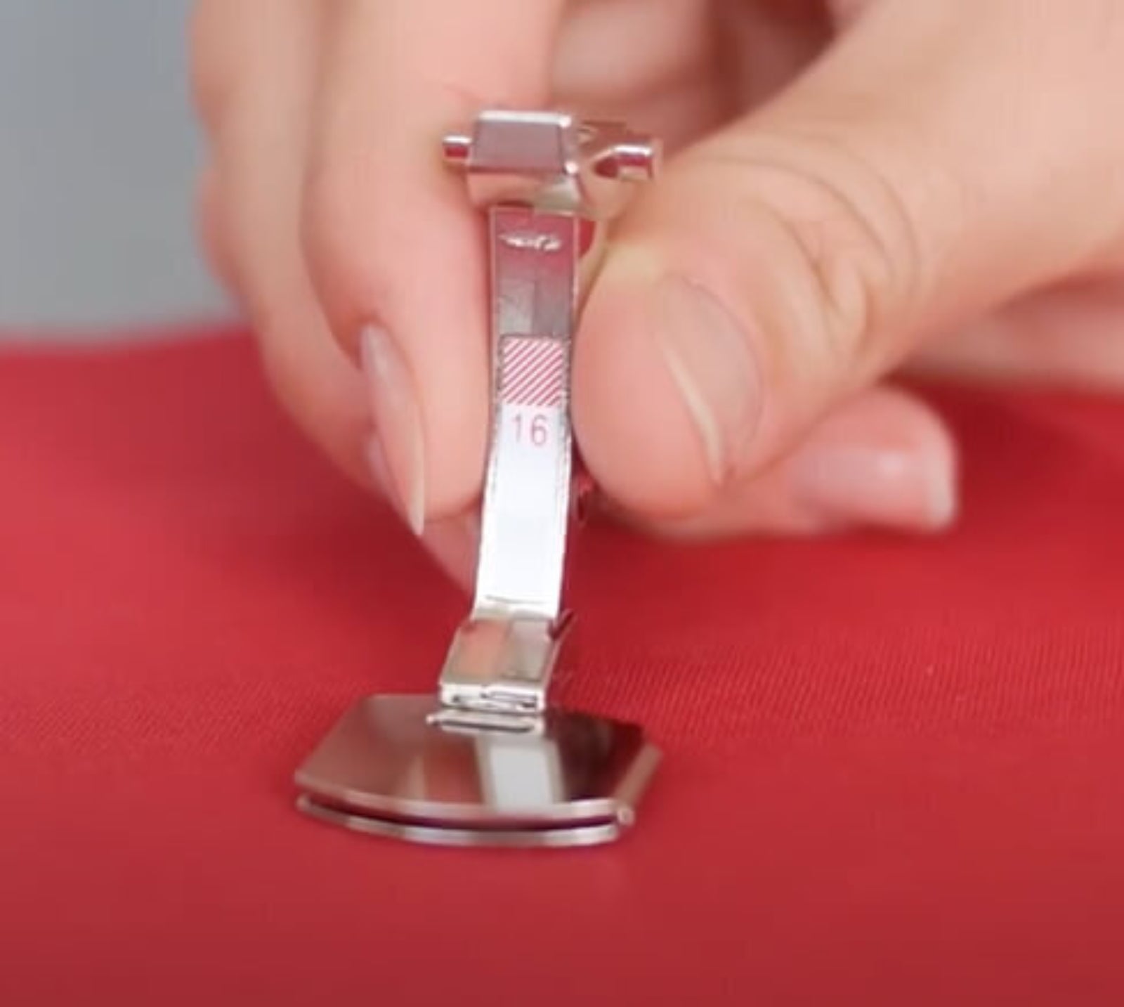 A close-up of a hand holding a silver sewing presser foot with #16 marking, positioned over red fabric.