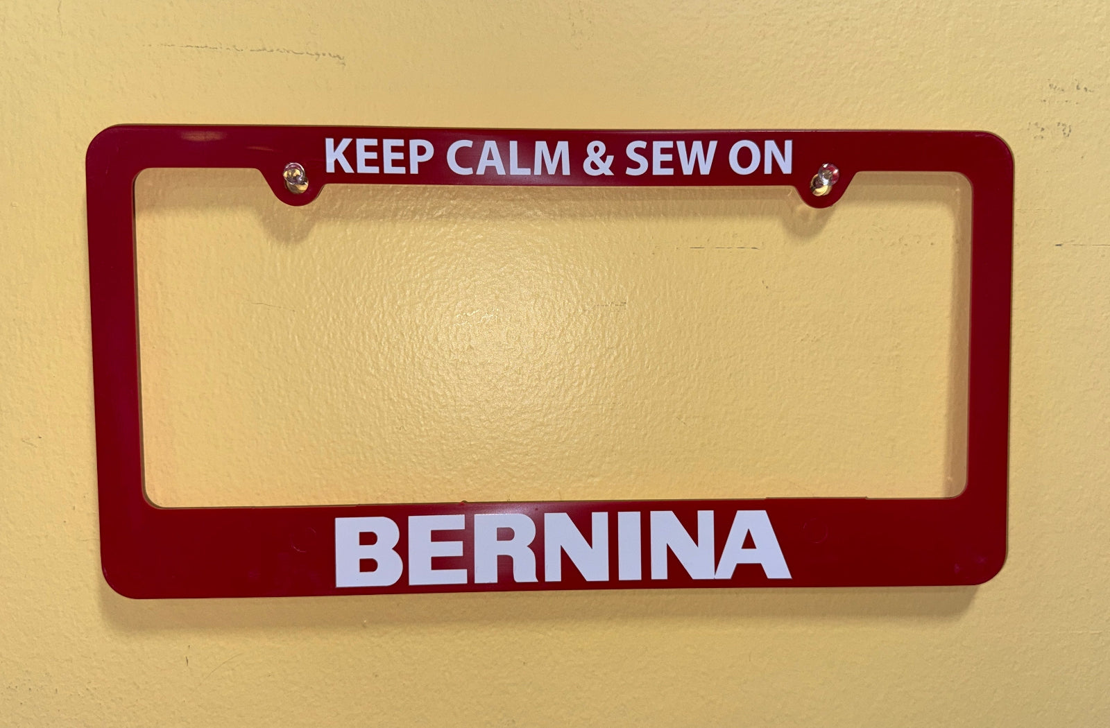 A dark red license plate frame with white text reading 'KEEP CALM & SEW ON' at the top and 'BERNINA' at the bottom, mounted against a light beige textured wall.