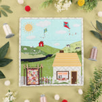 A square quilted fabric panel featuring a whimsical countryside scene with a house, barn, bicycle, flowers, hills, and kites, styled in a flat lay with thread spools, flowers, and leaves on a pale yellow background.