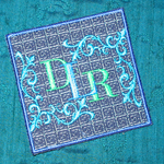 Embroidered monogram coaster with ornate blue scrollwork and green lettering on teal fabric.