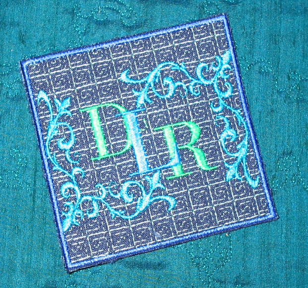 Embroidered monogram coaster with ornate blue scrollwork and green lettering on teal fabric.