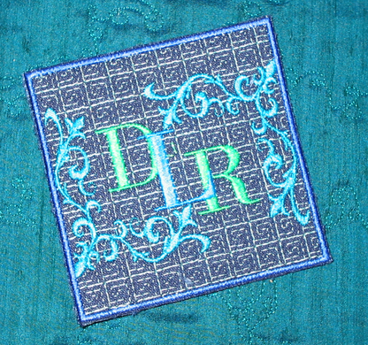 Embroidered monogram coaster with ornate blue scrollwork and green lettering on teal fabric.