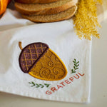 A white fabric napkin or kitchen towel with an embroidered acorn design and the word 'GRATEFUL' in red, styled with bread slices, a pumpkin, and dried yellow grass.