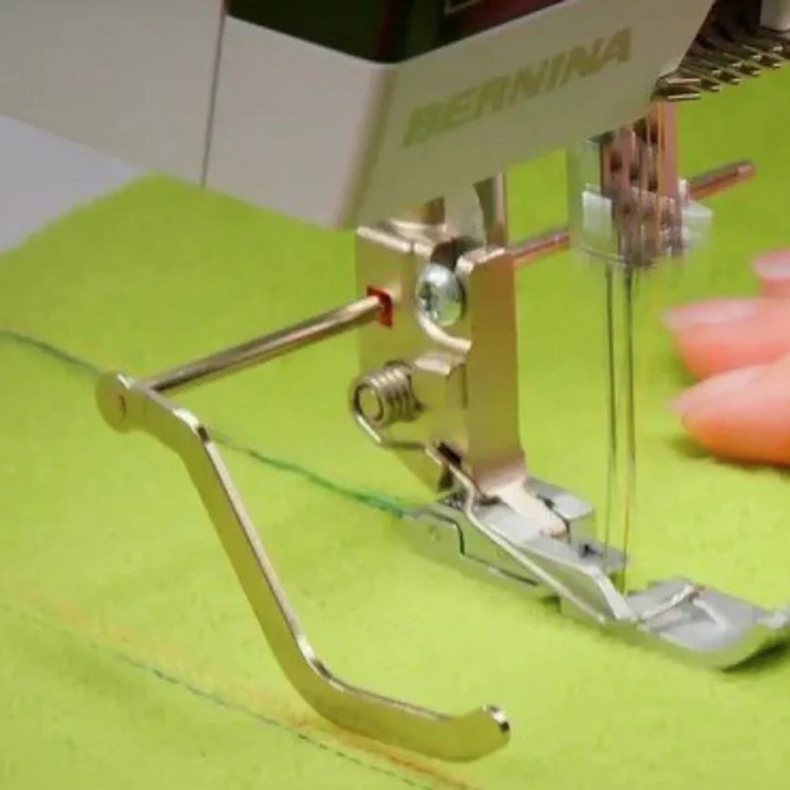 Close-up of a Bernina sewing machine with a curved metal presser foot stitching bright lime green fabric, with a finger visible on the right side.