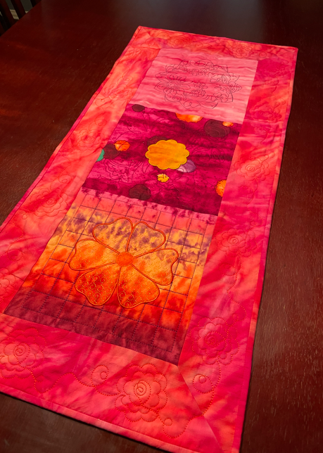 Quilted table runner in red, pink, and orange tones featuring floral motifs and decorative stitching.