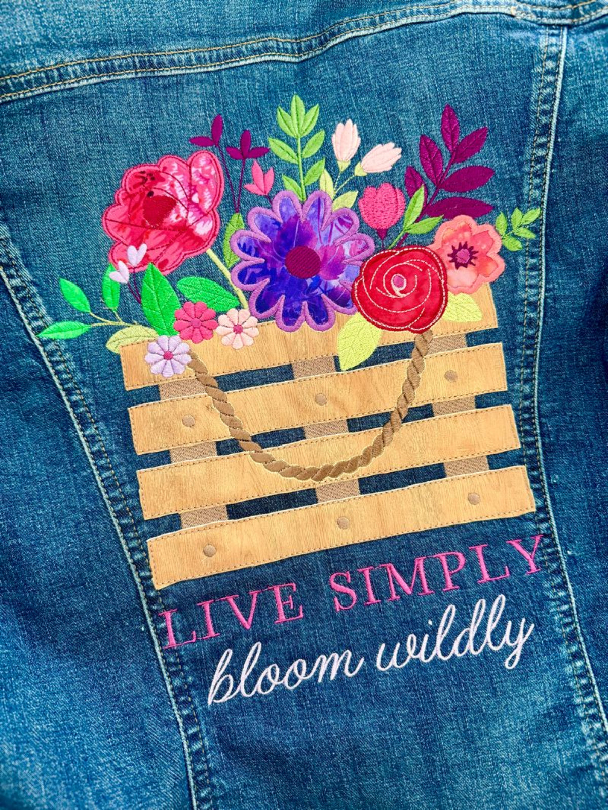 Close-up of a blue denim garment featuring a colorful embroidered basket of flowers and the text 'LIVE SIMPLY bloom wildly' on the back.