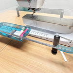A clear acrylic sewing machine guide with a blue and white measuring template attached, set up on a wooden table next to a sewing machine, with blue-handled scissors and an orange-handled tool nearby.