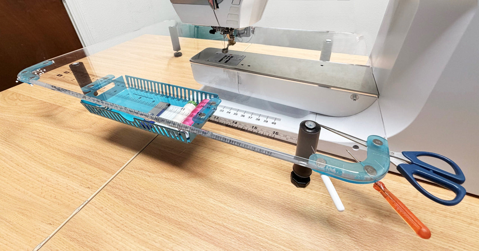A clear acrylic sewing machine guide with a blue and white measuring template attached, set up on a wooden table next to a sewing machine, with blue-handled scissors and an orange-handled tool nearby.